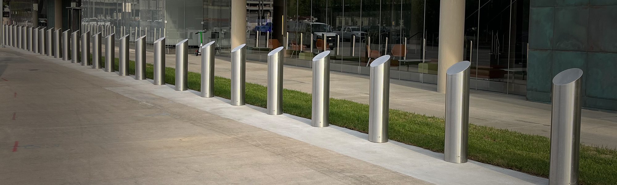 Row of brushed stainless steel slope-top bollards lining a glass office building plaza